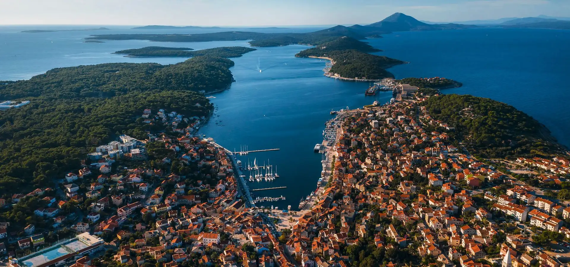 An aerial view of Mali Lošinj, Croatia, showcasing the vibrant coastal town nestled around a deep, sheltered bay filled with sailboats. Lush green islands and the open sea stretch into the distance under a clear sky.