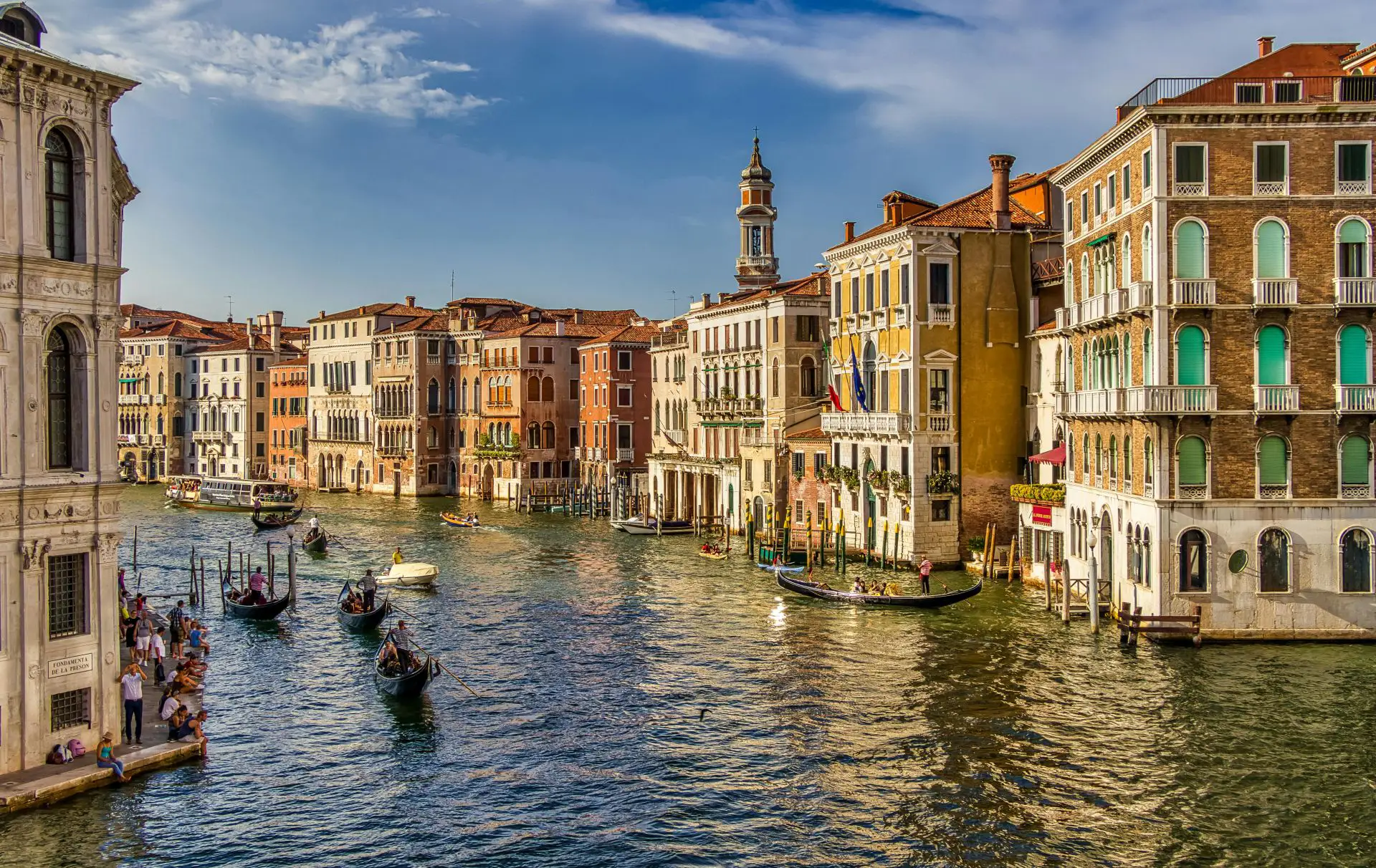 A scenic view of the Grand Canal in Venice, Italy, with numerous gondolas and boats traversing the water. Historic multi-story buildings with colorful facades line both sides of the canal under a partly cloudy blue sky.
