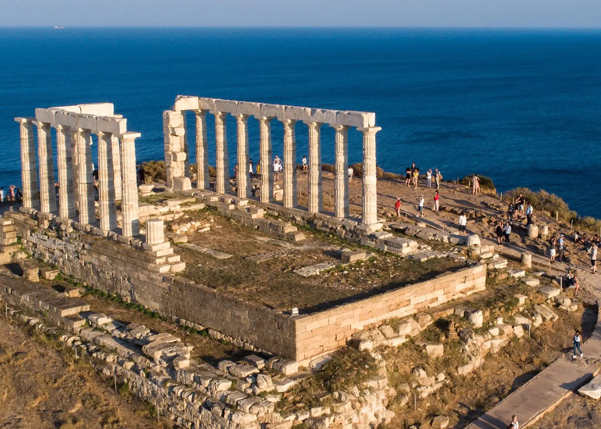 High-angle photograph of the Temple of Poseidon ruins at Cape Sounion, displaying ancient white Doric columns atop a rocky promontory overlooking the deep blue Aegean Sea.