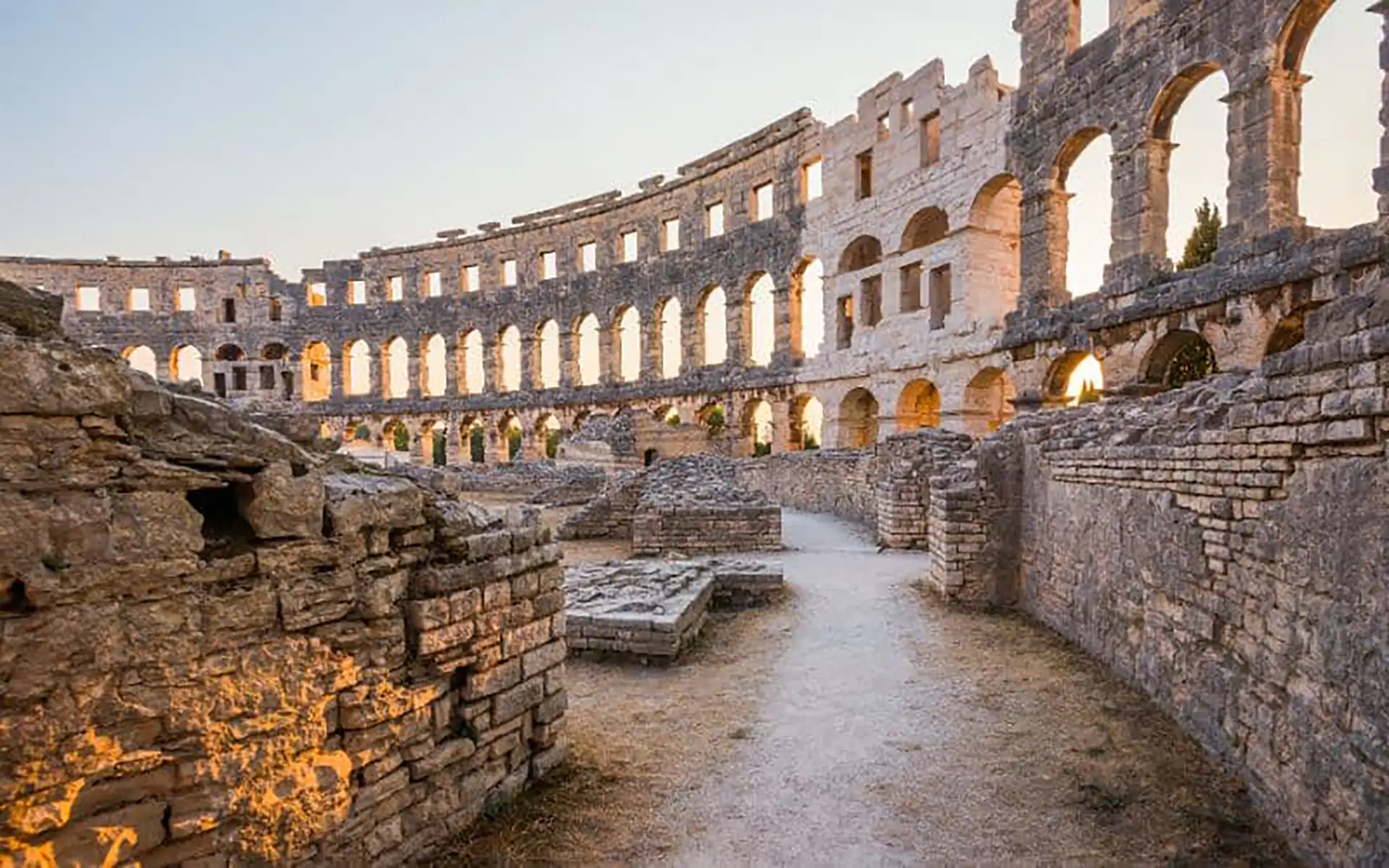 Copyright: kaycco//Adobe Stock Interior view of the Pula Arena, a well-preserved Roman amphitheater in Pula, Croatia. Sunlight streams through arched openings along the upper walls, illuminating the stone structure.