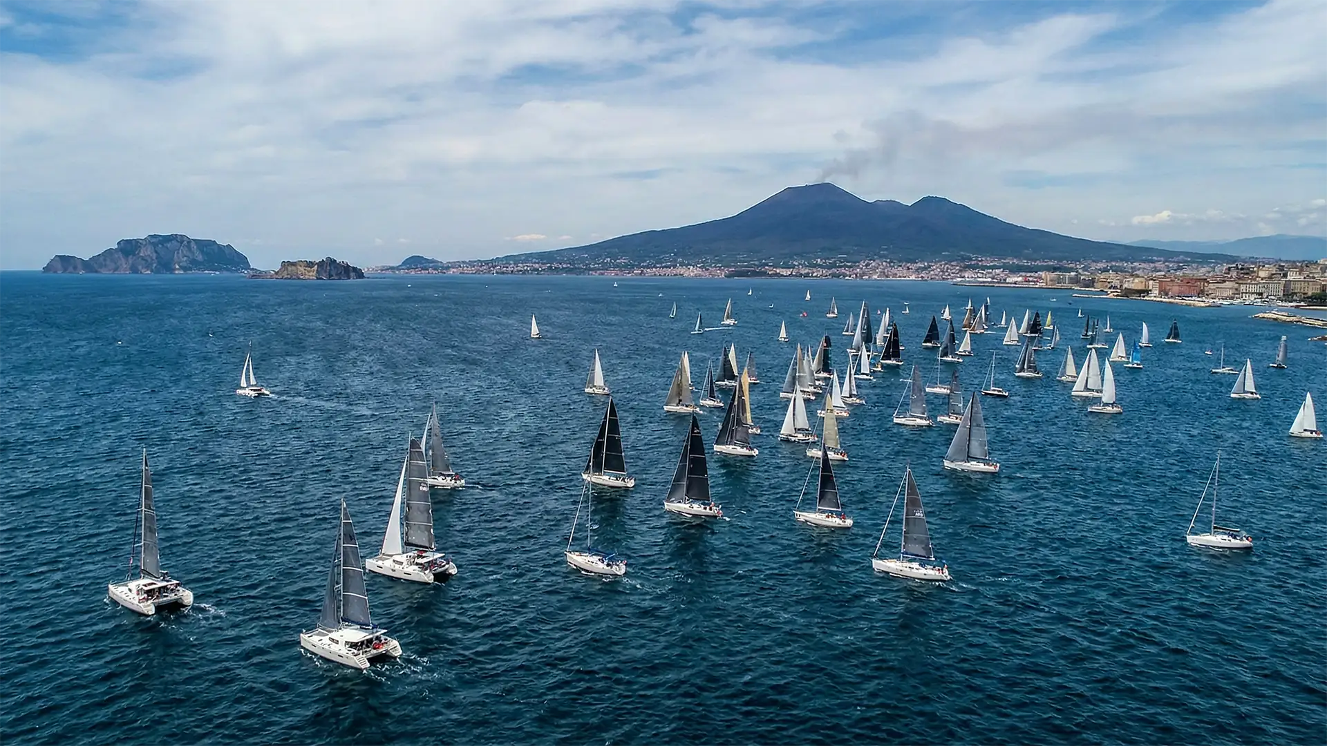 A wide aerial view of the Bay of Naples on a sunny day, showing dozens of sailboats with white and black sails scattered across the deep blue water, with the coastline, the island of Capri on the left, and Mount Vesuvius emitting a faint plume of smoke in the background.