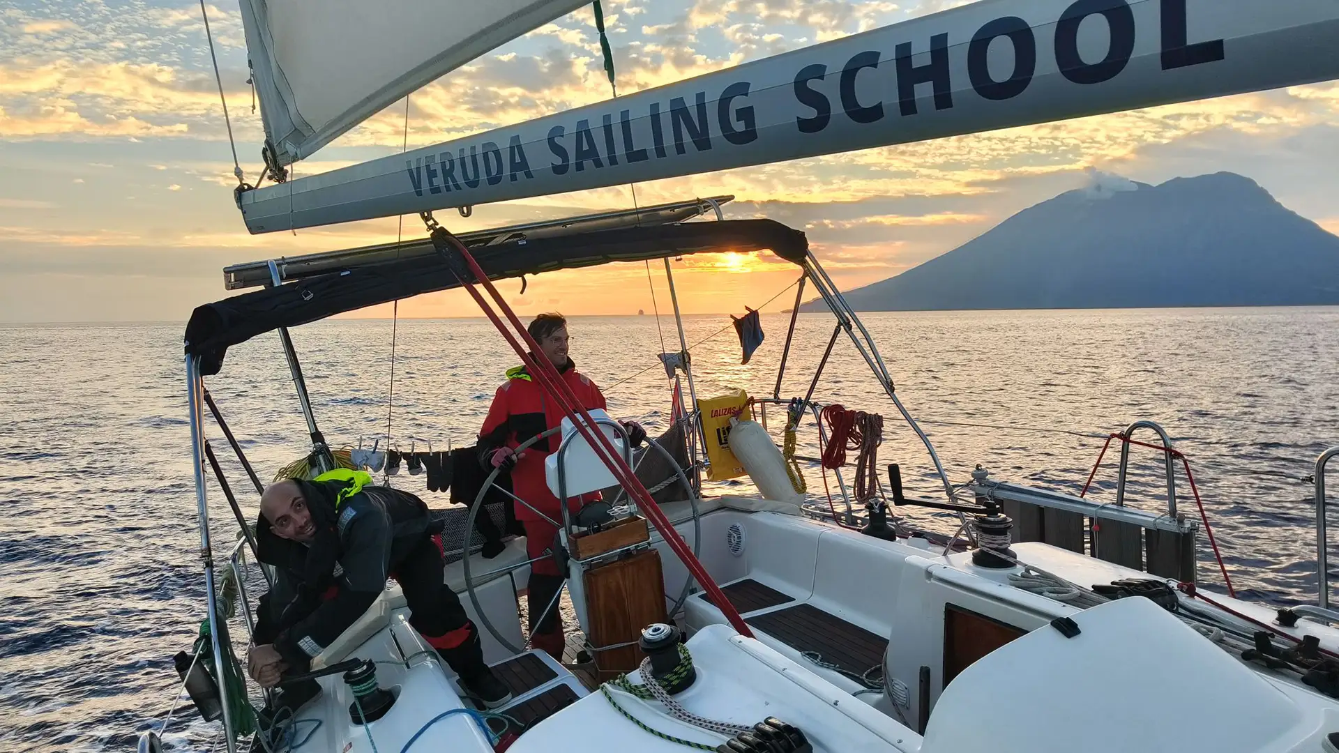 A racing crew on an offshore sailboat. Behind the boat, the large volcanic cone of Stromboli volcano is visible, spewing clouds of steam.
